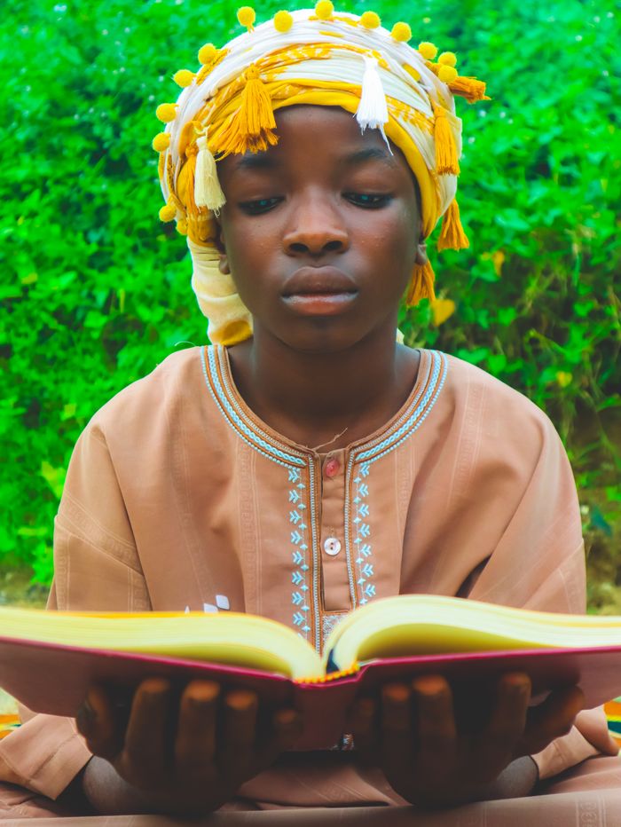 A focused African child wearing traditional clothing reads a book outdoors.