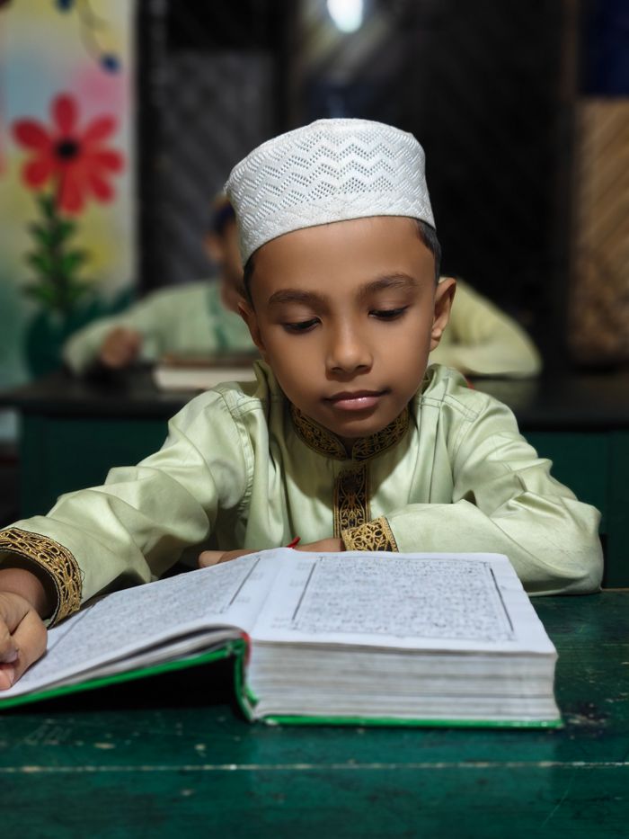 A young boy in traditional attire reads the Quran attentively at a classroom desk.