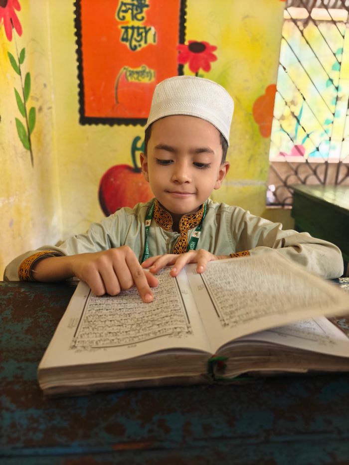 A young boy in traditional attire reads a religious book indoors, focused intently.