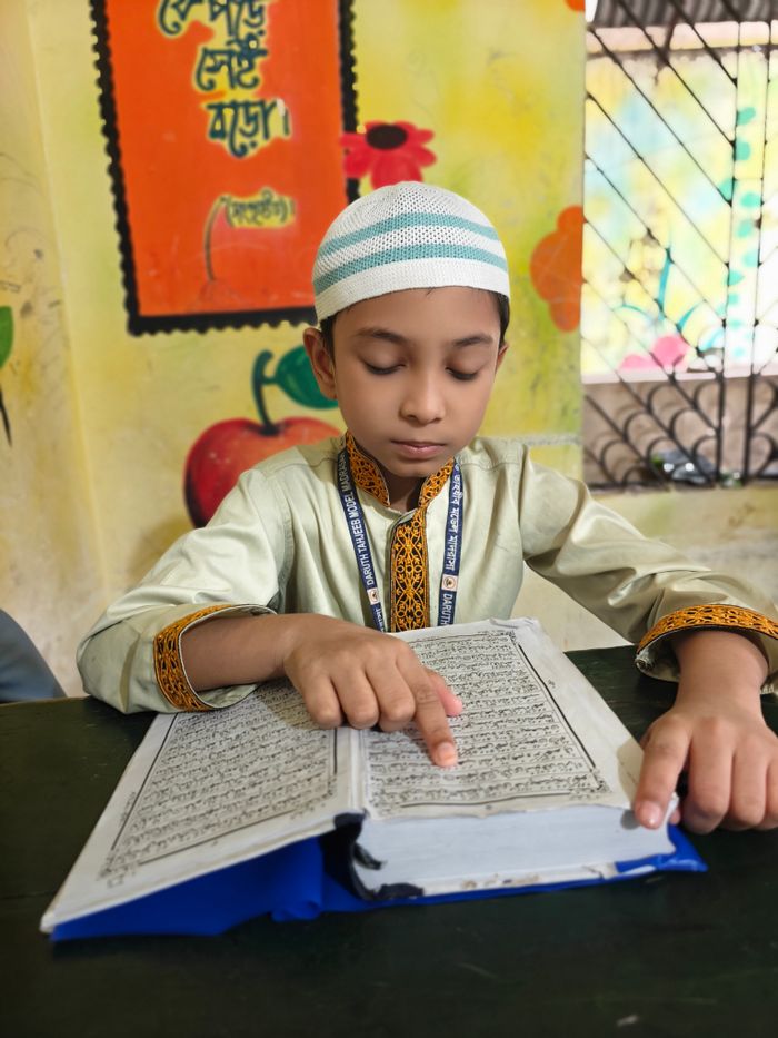 A young boy in traditional attire reading a religious book inside a classroom.
